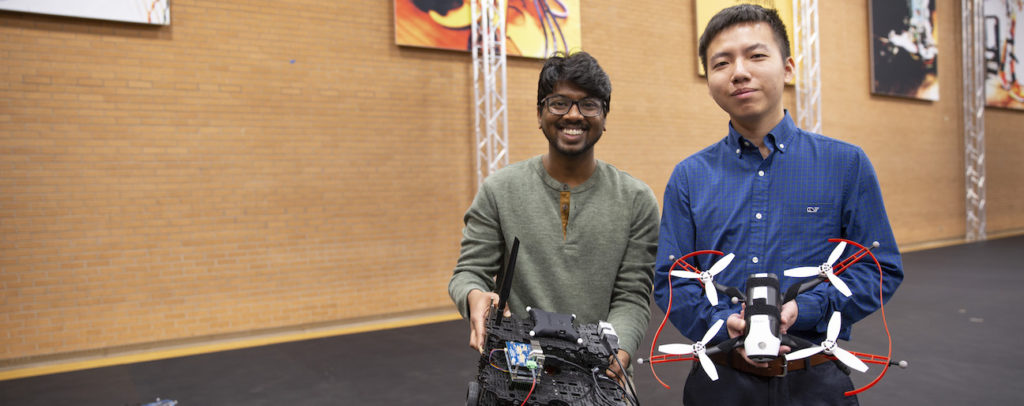 Two students smiling and displaying a small robot and a drone